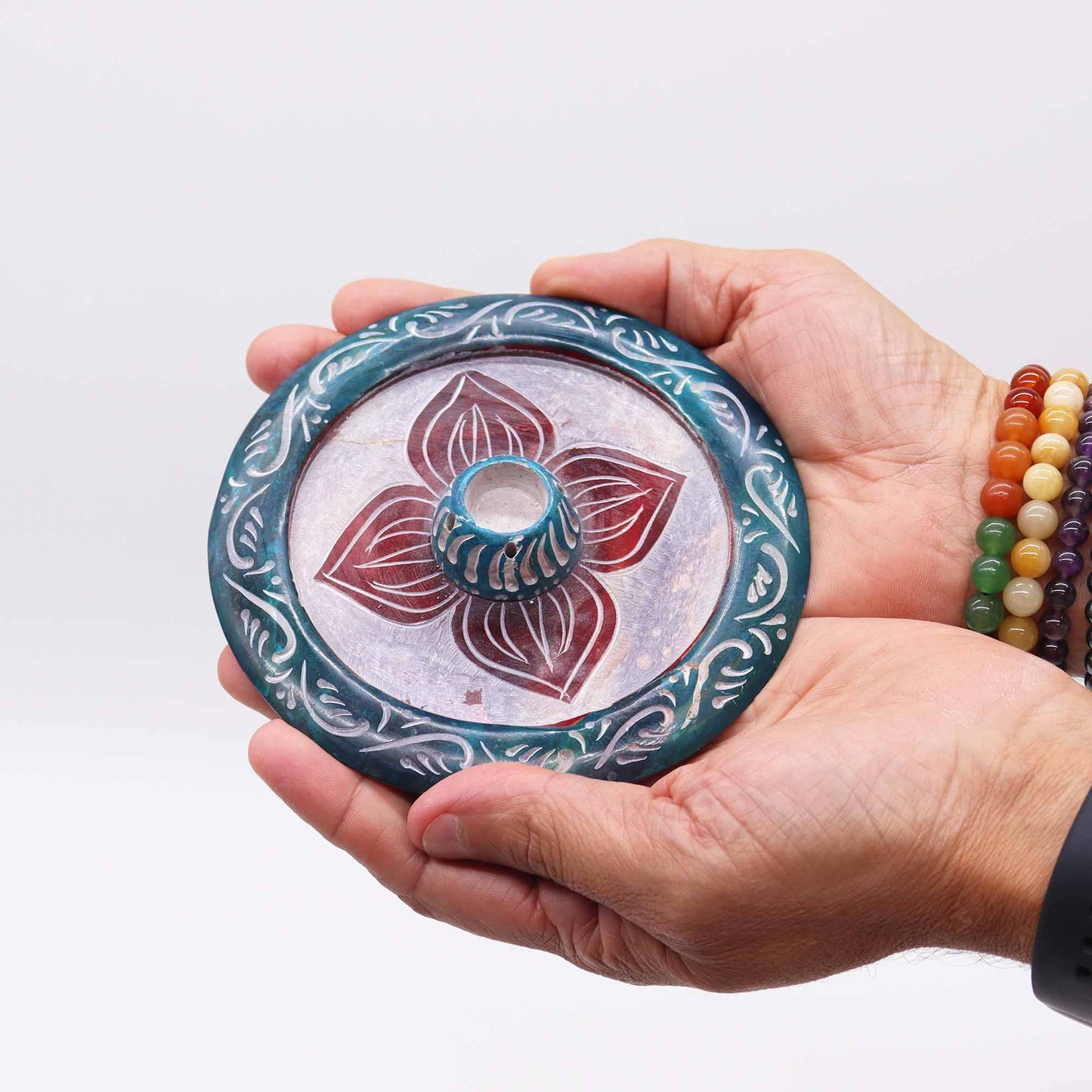 Round soapstone incense holder with carved four-petal flower design, dyed in teal and burgundy, holds both incense cones and sticks.