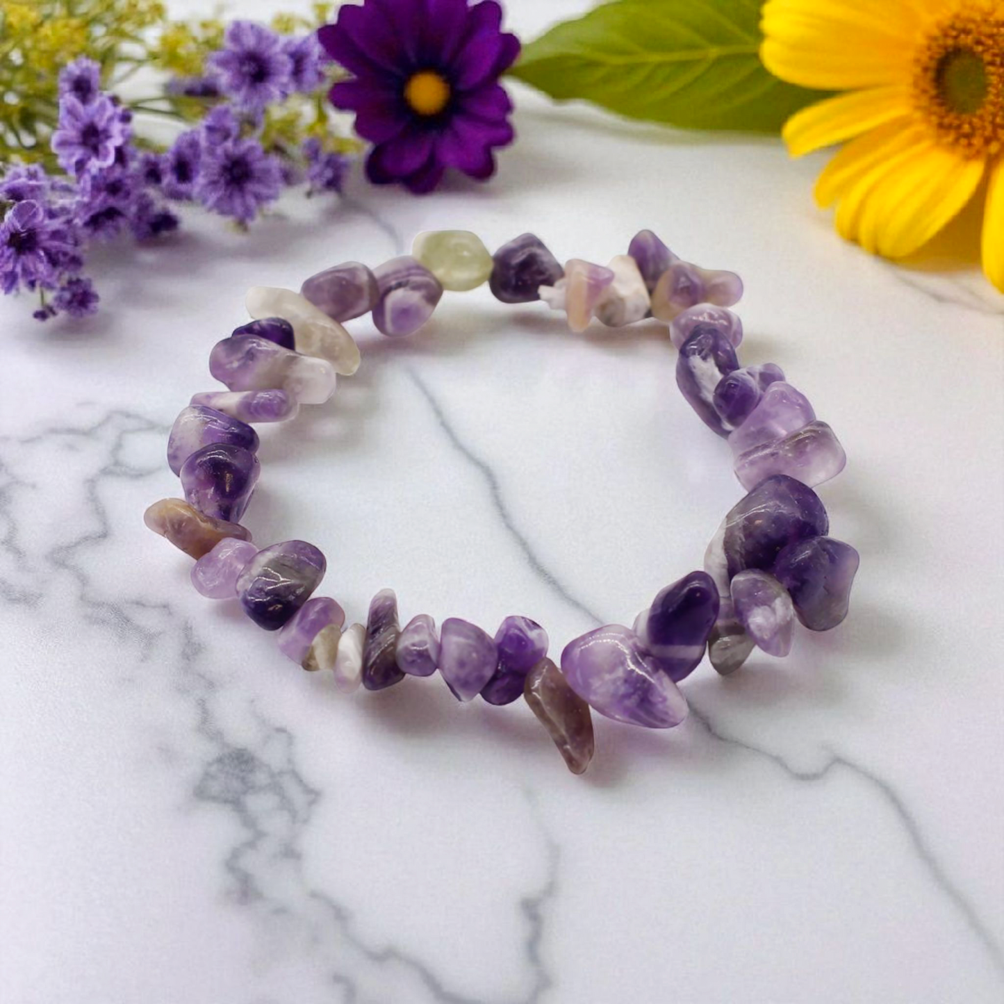 Amethyst chip stone bracelet with soft purple stones, on a white marble table top with wild flowers in the background, symbolising calm, clarity, and spiritual awareness.