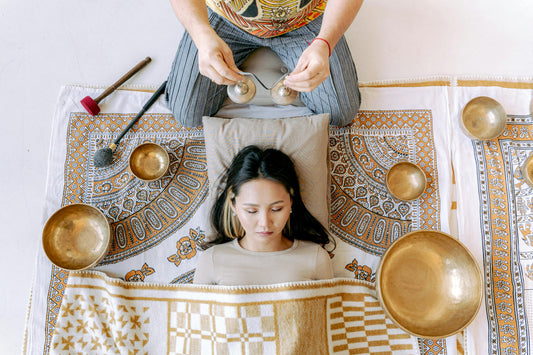 Woman laying down on an ornate rug with here eyes closed while a sound healing practitioner is above her with cymbals, Tibetan singing bowls of different sizes all around the woman 