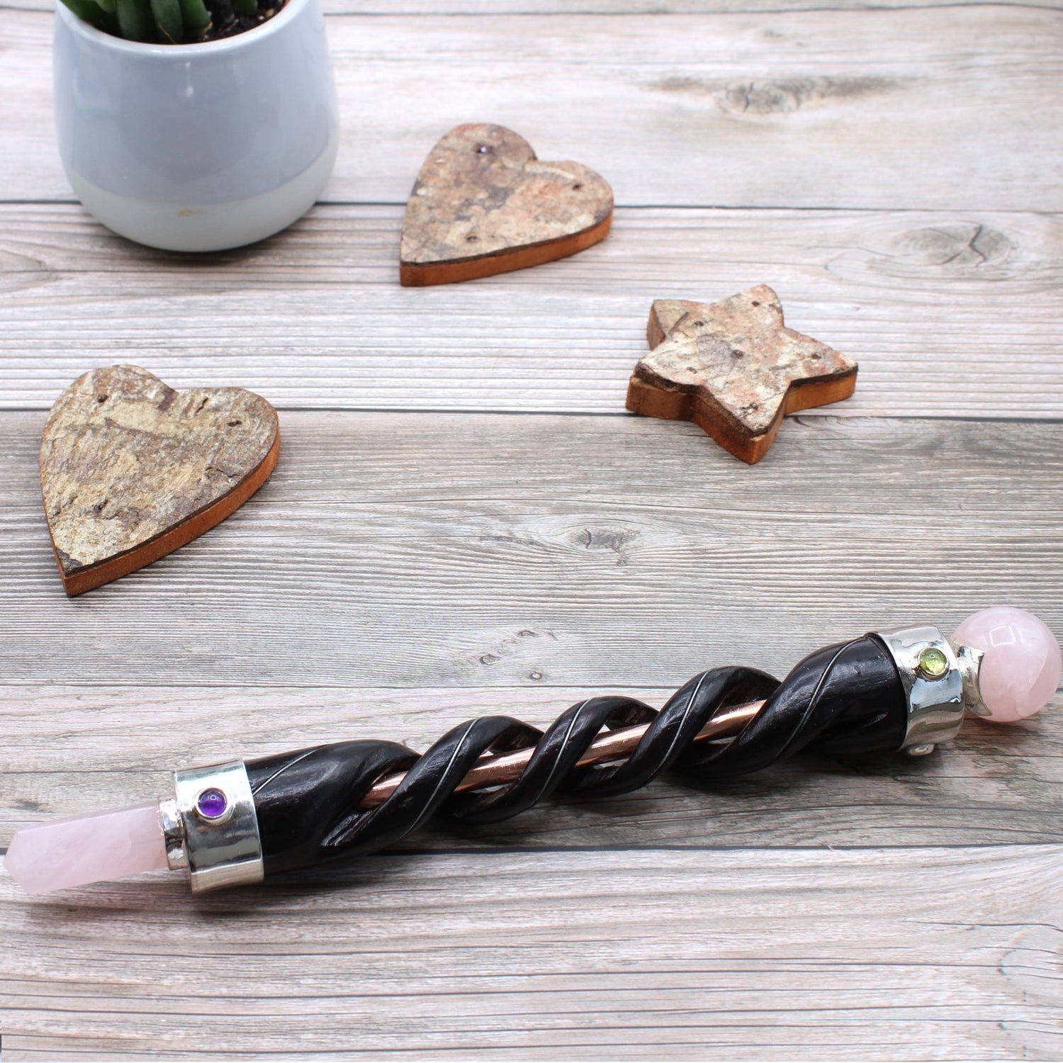 Crystal wand on a table, wand made from twisted dark coloured wood, copper tube and silver plated copper, contain an orb rose quartz crystal on one end and a pointed one on the other