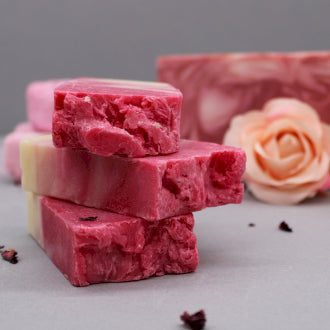 Stack of three bars of soap pink in colour with dried rose petals on the ground and a whole rose bud in the background 