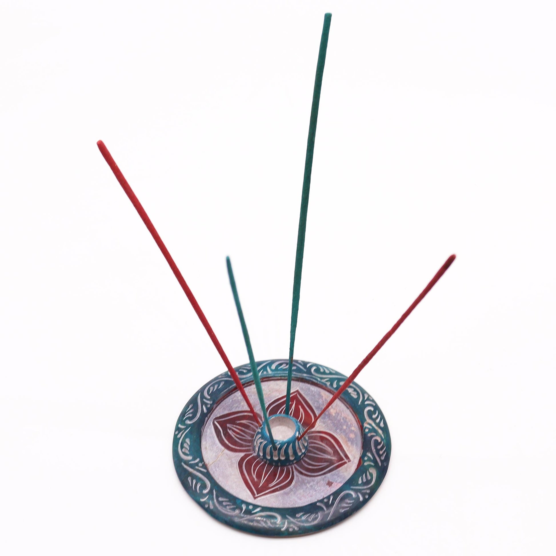 Round soapstone incense holder with carved four-petal flower design, dyed in teal and burgundy, holds both incense cones and sticks.

