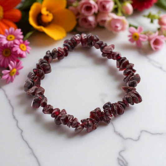 Blood Garnet crystal chip stone bracelet with deep red stones, on white marble table top with wild flowers in the background, symbolising strength, passion, and emotional resilience.
