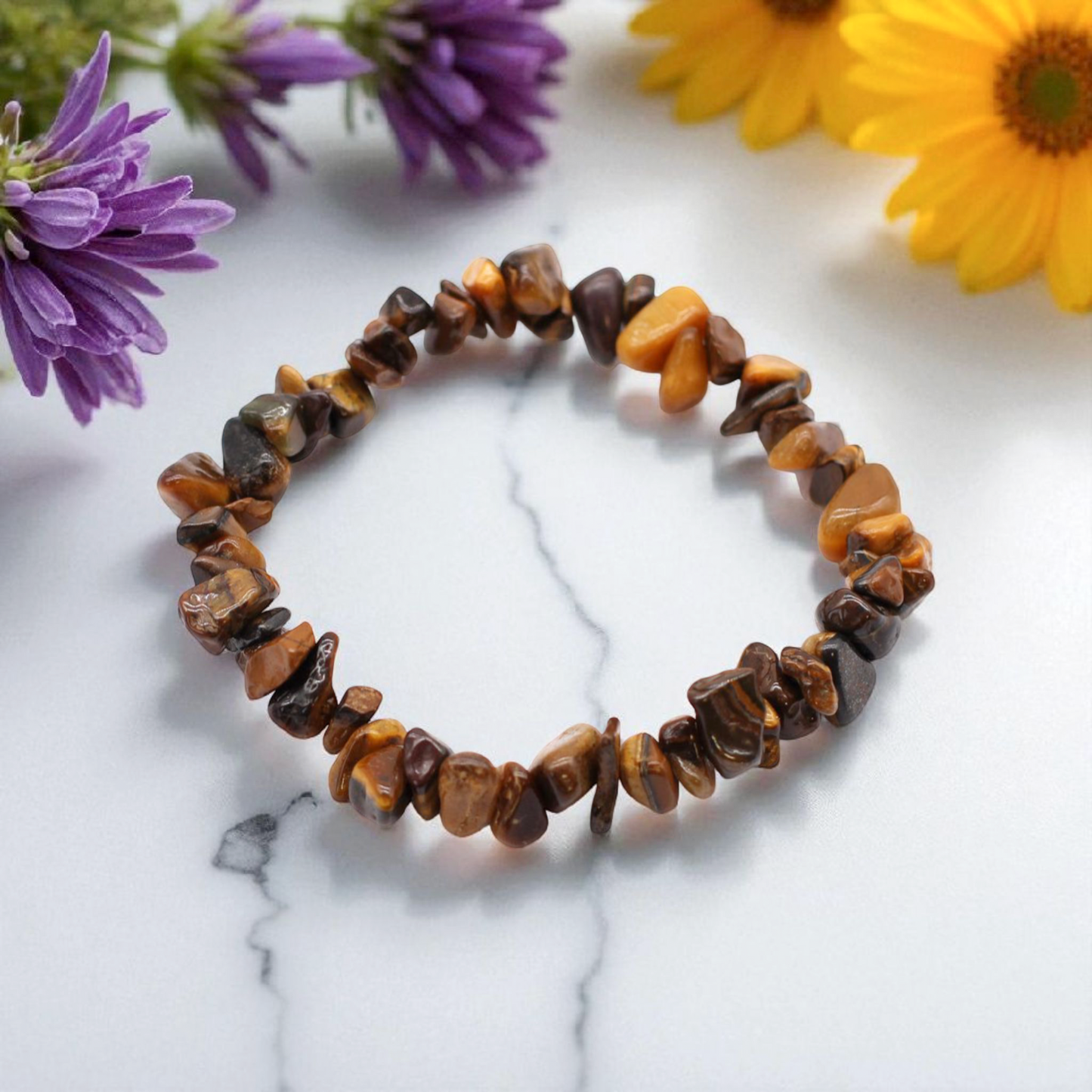 Tiger’s Eye chip stone bracelet with golden-brown stones, on a white marble table top with wild flowers in the background, symbolising confidence, protection, and grounded strength.
