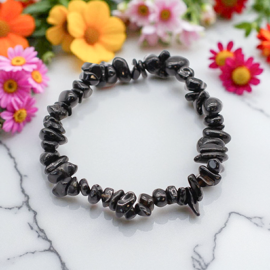 Black Agate chip stone bracelet with deep black stones, on a white marble table top with wild flowers in the background, symbolising protection, grounding, and emotional resilience.
