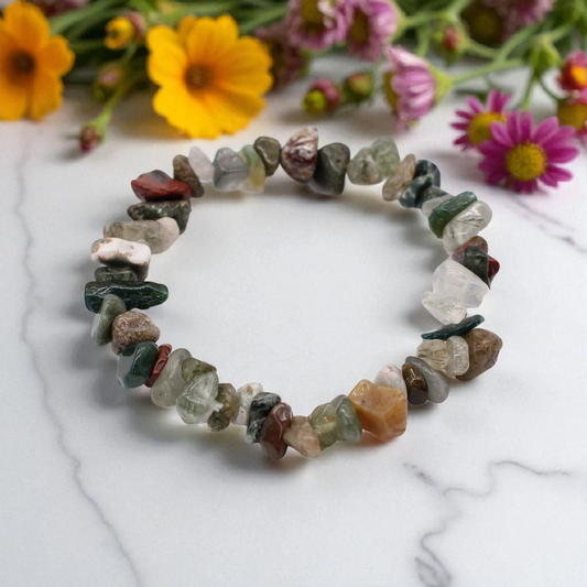 Moss Agate chip stone bracelet with green marbled stones, on a white marble table top with wild flowers in the background, symbolising balance, renewal, and connection to nature.
