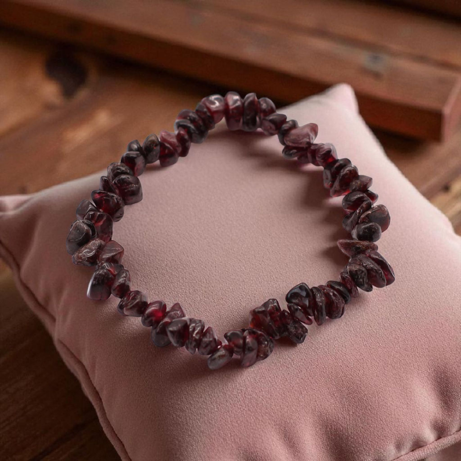 Blood Garnet crystal chip stone bracelet with deep red stones, on a pillow on a wood table, symbolising strength, passion, and emotional resilience.
