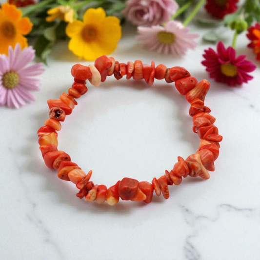 Chip stone bracelet made with redish pink Coralite stones, on white marble table top with wildflowers in the background, symbolising vitality, emotional healing, and inner peace.
