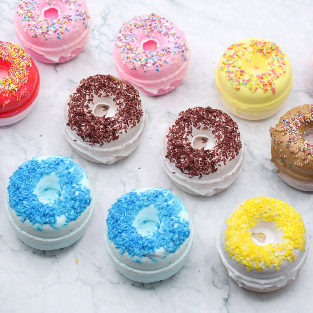 Collection of bath bombs, all in the shape of donuts and all with sprinkles on top, various colours from pink, yellow and white.